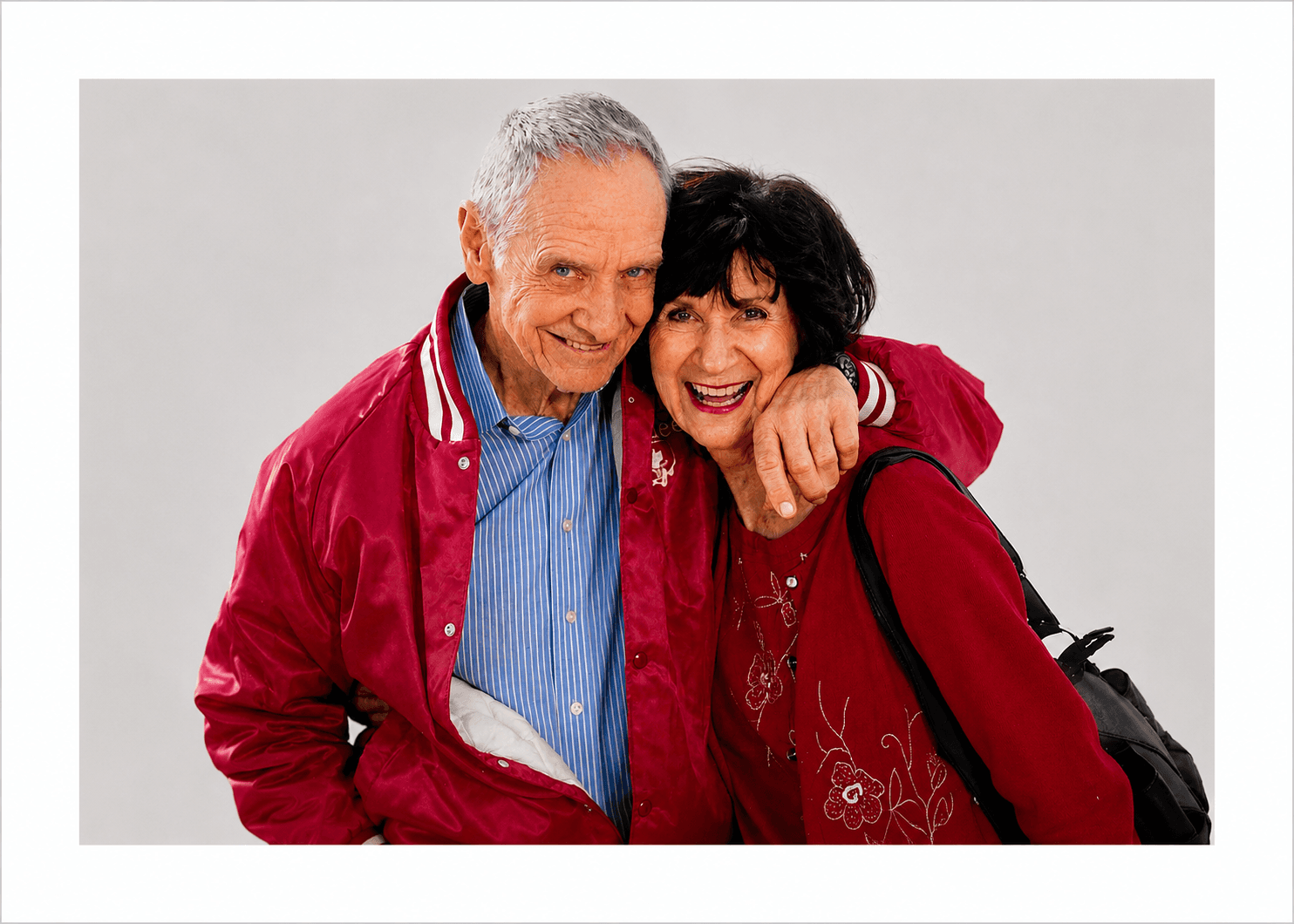Smiling elderly man and woman in red outfits hugging against a plain grey background.